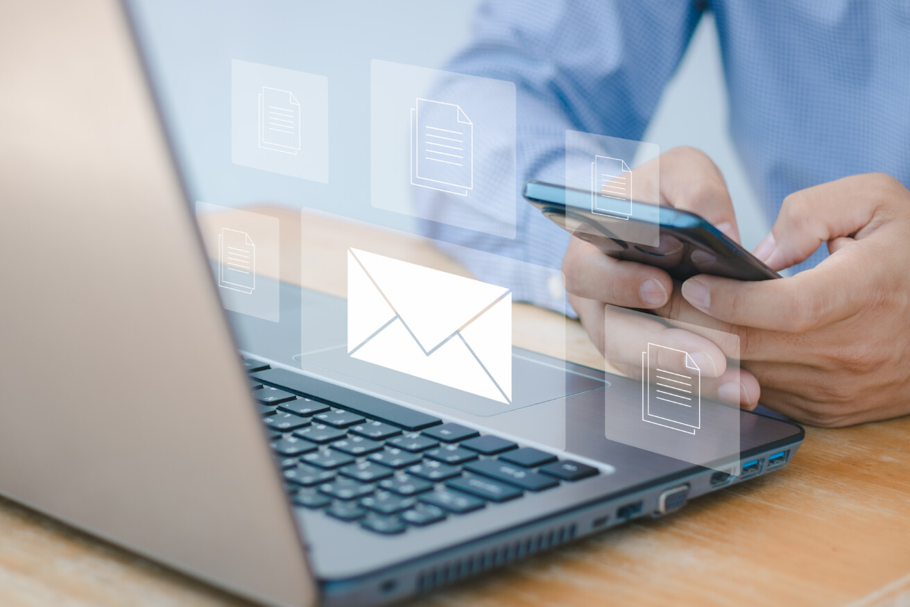 A person uses a smartphone and laptop at a desk, with digital icons of an envelope and documents floating above the devices, symbolizing email and file sharing.