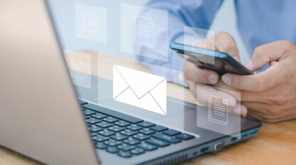 A person uses a smartphone and laptop at a desk, with digital icons of an envelope and documents floating above the devices, symbolizing email and file sharing.