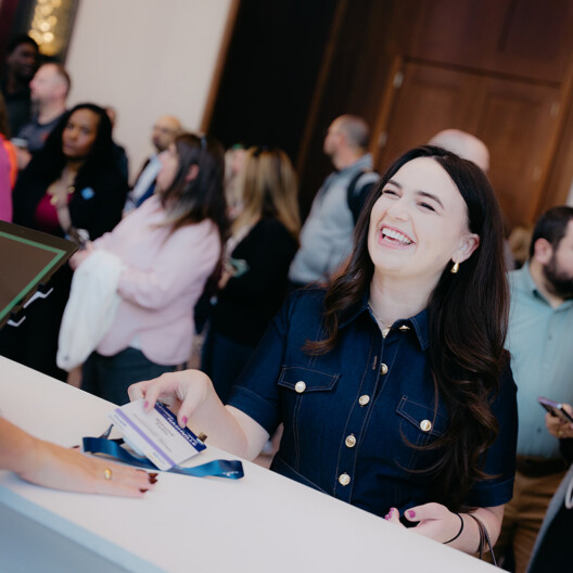 A woman smiles while checking in at a registration desk, handing over a badge, with a group of people waiting in line behind her.