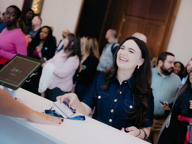 A woman smiles while checking in at a registration desk, handing over a badge, with a group of people waiting in line behind her.