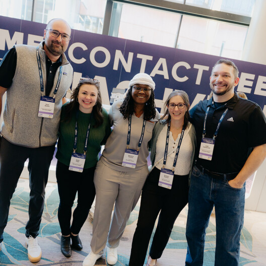 Five people stand and smile for a group photo at an event check-in area, wearing name badges. A check-in counter and event signage are visible in the background.