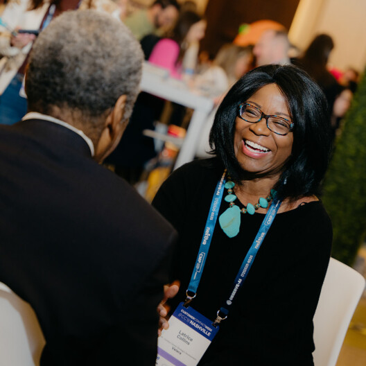 Two people sit and talk at a professional event; the woman in focus is smiling, wearing glasses, a blue necklace, and a name badge. Others are visible in the background.