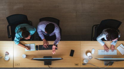 Three people working at computers at a shared wooden desk, seen from above; one person writes in a notebook, another gestures, and the third types on a keyboard.