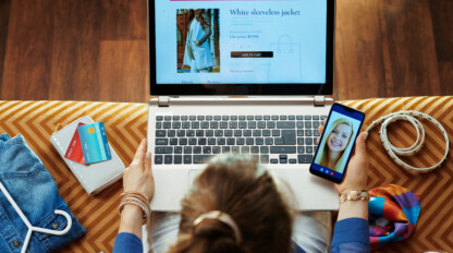A person shops for clothes online on a laptop while video chatting on a smartphone; credit cards, a belt, and clothes are nearby on the sofa.