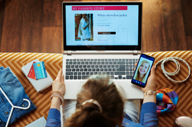 A person shops for clothes online on a laptop while video chatting on a smartphone; credit cards, a belt, and clothes are nearby on the sofa.