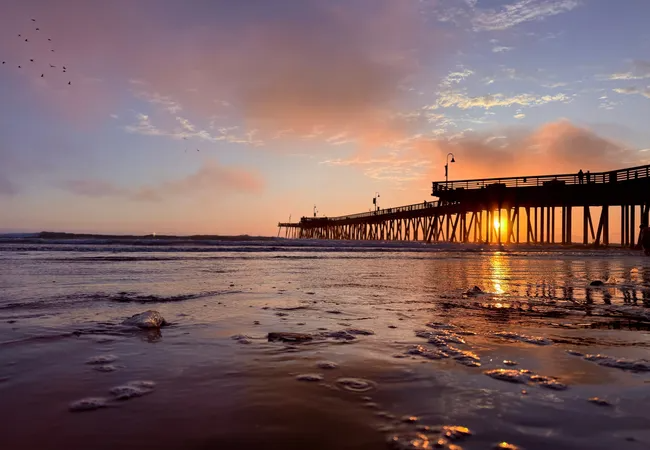 A low-angle, wide shot of the Pismo Beach Pier at sunset. The pier’s silhouette stretches across the mid-ground, with the sun glowing brightly between its support pilings. In the foreground, wet sand and shallow ocean water reflect the vibrant orange and purple hues of the sky. Fluffy, pink-tinted clouds fill the sky, and a small flock of birds is visible in the upper left corner.