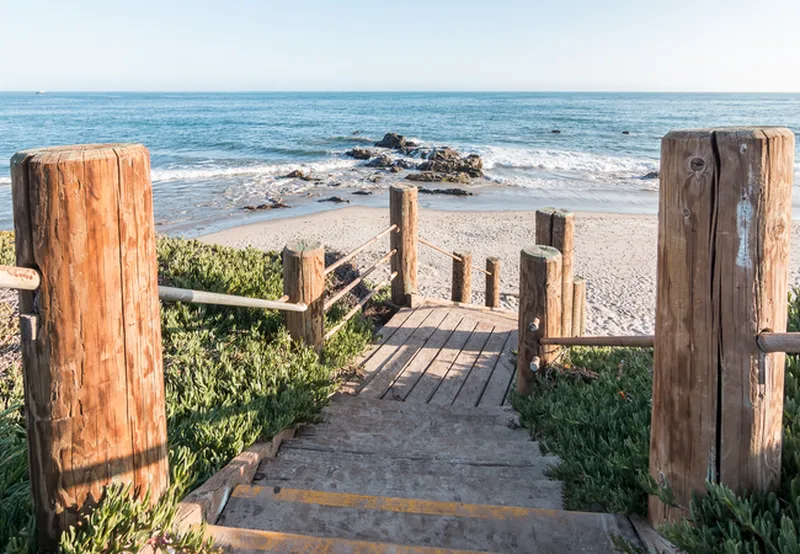 A first-person perspective looking down a set of wooden and concrete stairs leading to a sandy beach. The stairs are flanked by thick wooden pilings connected by rope railings and surrounded by green coastal ice plant. In the background, small rocky outcrops sit in the turquoise ocean water under a bright, clear sky.