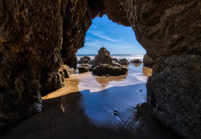 A view from inside a dark, jagged sea cave looking out toward the bright Pacific Ocean. The dark, textured rock walls of the cave frame a natural arch. Through the opening, several large sea stacks and boulders sit on a wet sandy beach, reflecting the bright blue sky and scattered white clouds above. The sunlight illuminates a patch of sand just outside the cave's mouth, creating a high-contrast, dramatic scene.