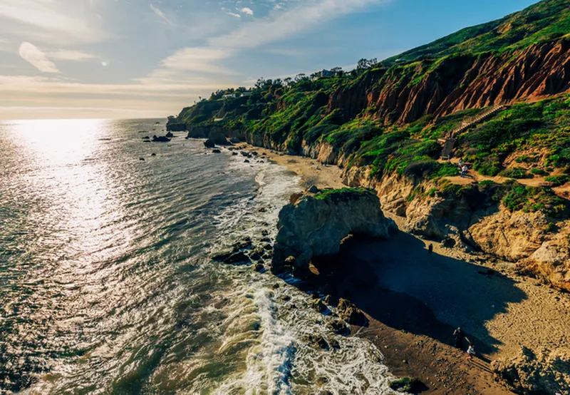 A stunning high-angle aerial view of a rugged coastline at sunset. On the right, steep, jagged cliffs covered in vibrant green foliage and brown earth drop down to a narrow sandy beach. In the center, a massive rock formation with a natural arch sits on the sand as white waves crash against it. The glittering ocean stretches to the left under a soft, hazy sky with the sun low on the horizon.