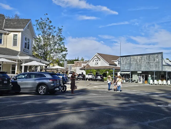 A wide-angle view of a sunlit street in a small town. Several two-story buildings with light-colored siding and dark grey roofs line the roadway, including one with a white porch and umbrellas. People are seen crossing the street and walking along the sidewalks, with parked cars including a silver SUV in the foreground under a bright blue sky with wispy white clouds.