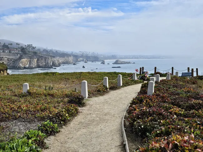 A high-angle, wide shot of a dirt walking path winding through low, reddish-green coastal shrubs. The path is lined with grey concrete posts and rope railings. In the background, rugged sea cliffs overlook a calm blue ocean, with several rocky sea stacks jutting from the water under a soft, hazy sky.