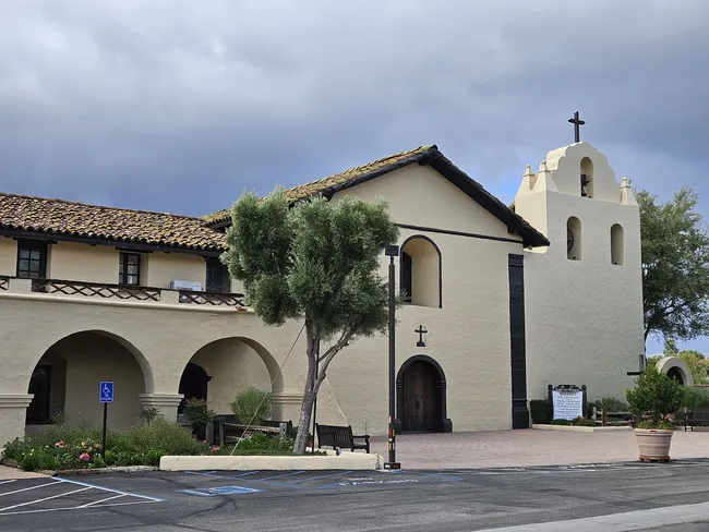 A wide-angle shot of a historic mission-style building with cream-colored stucco walls and a weathered brown tile roof. On the left, an arched walkway leads into a courtyard, and on the right, a white bell tower topped with a small black cross rises under a cloudy grey sky. A small, bushy green tree and several park benches are in the foreground, bordering a paved parking lot.