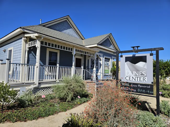 A wide-angle view of a light blue, single-story house with white trim and a small front porch featuring white railings. A large grey and white sign stands in the foreground to the right, displaying text and a graphic of a sand dune. A paved path leads to the house through a small garden filled with green shrubs and red-flowering plants under a clear, bright blue sky.