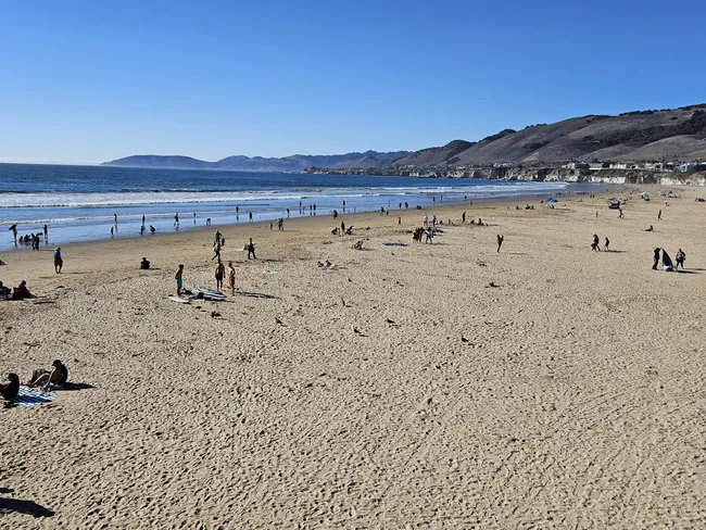 A high-angle, wide-angle shot of a broad, sandy beach bustling with people on a sunny day. Tiny figures of beachgoers are scattered across the sand and along the shoreline where gentle waves meet the coast. In the background, dark, rolling hills and distant coastal mountains stretch along the deep blue ocean under a vast, clear sky.