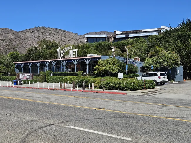 A wide-angle shot of a classic roadside restaurant called "Neptune's Net." The building is a single-story, blue-grey structure with a large, white neon sign on the roof. It sits along a paved highway, with a white SUV parked nearby. Dense green trees and brush surround the building, with a massive, rocky brown mountain rising directly behind it under a clear blue sky.