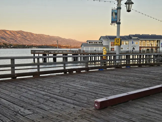 A low-angle view of a wide wooden pier with weathered planks and heavy timber railings. In the mid-ground, a light grey building with solar panels and a "10 MPH" sign sits on the pier. In the background, calm ocean water reflects a soft sunset glow, framed by a massive range of rolling brown mountains under a pale evening sky.