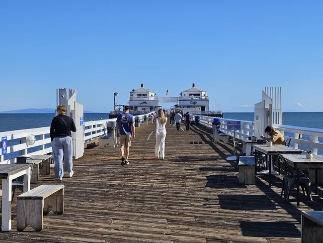 A perspective view looking down a long, weathered wooden pier stretching over a deep blue ocean. People are seen strolling along the pier, while others sit at small outdoor tables and benches on either side. At the end of the pier stand two small, symmetrical white buildings with dark roofs, all under a vast, cloudless blue sky with a distant hazy mountain visible on the horizon.