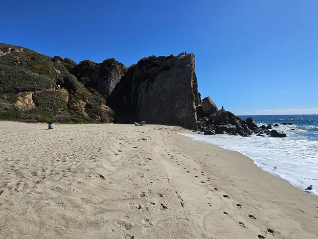 A low-angle view of a sandy beach meeting a massive, dark rock cliff that rises sharply on the right. Gentle white surf washes over the sand in the foreground, while a steep, green-covered hillside rises to the left. A clear, deep blue sky stretches above the coastline, and a few small figures are visible near the base of the rocks.