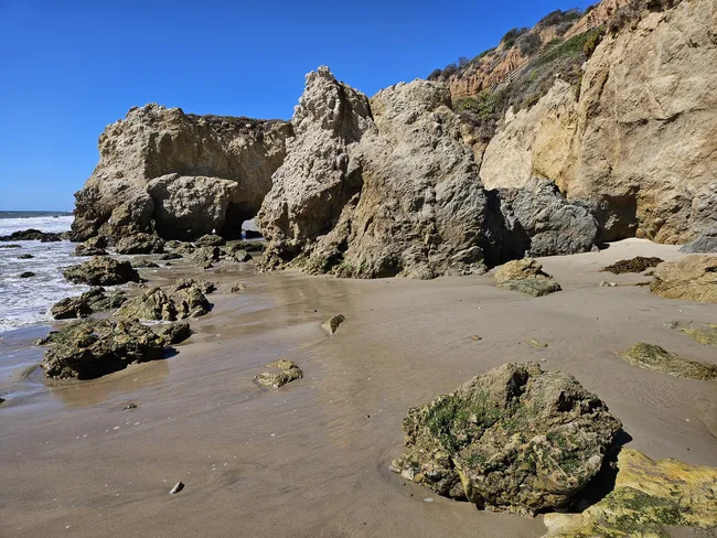A low-angle view of a sandy beach meeting a massive, dark rock cliff that rises sharply on the right. Gentle white surf washes over the sand in the foreground, while a steep, green-covered hillside rises to the left. A clear, deep blue sky stretches above the coastline, and a few small figures are visible near the base of the rocks.