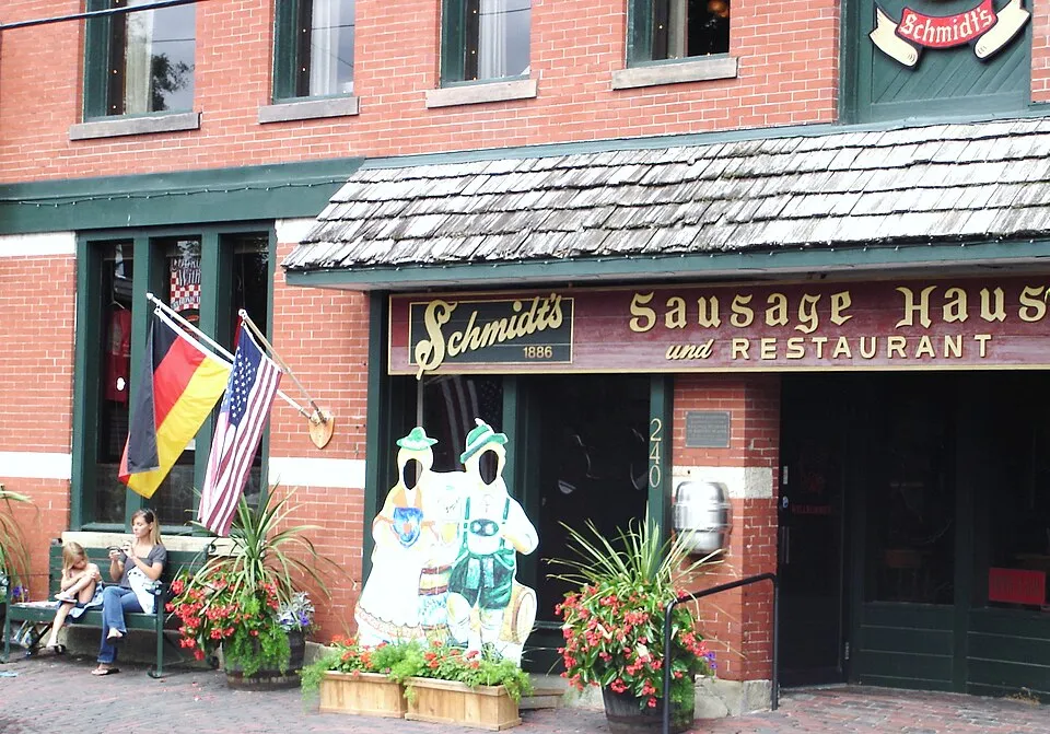 Exterior of Schmidt's Sausage Haus und Restaurant, a historic German-American eatery established in 1886 in German Village, Columbus, Ohio. The red brick building features a wood-shingled awning with gold lettering reading "Schmidt's Sausage Haus und Restaurant." An American flag and a German flag are mounted side by side near the entrance. A face-cutout photo prop depicting two figures dressed in traditional Bavarian attire stands out front, flanked by potted red flowers and tropical plants. Two women are seated on a bench to the left.