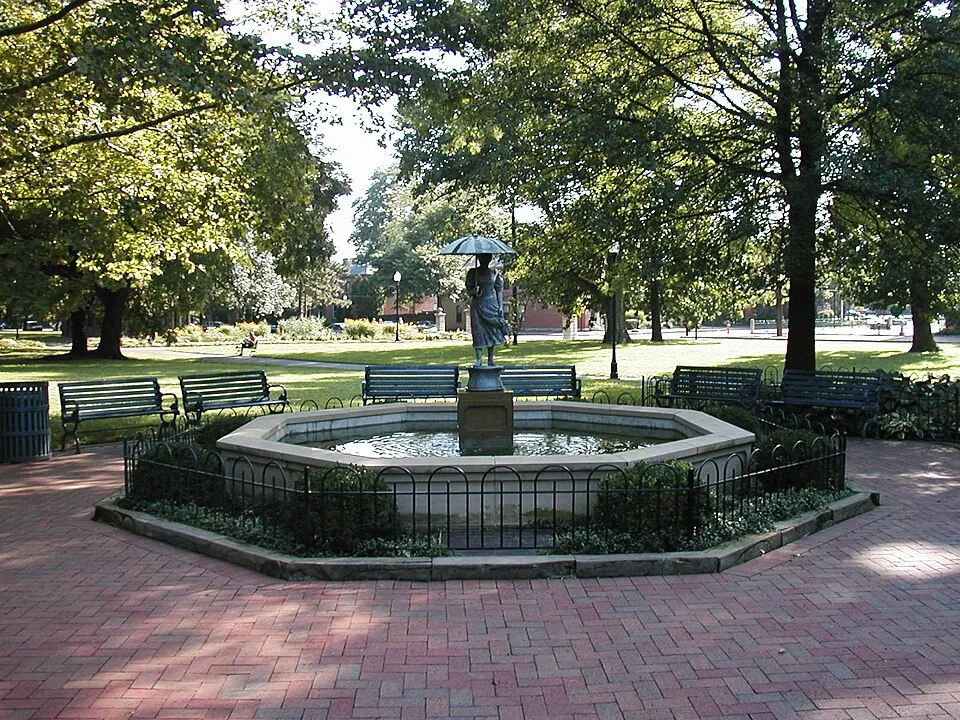 A bronze statue of a woman holding an umbrella stands at the center of an octagonal stone fountain pool, surrounded by a low black iron fence and low plantings. The fountain is set on a red brick plaza encircled by several green park benches. Large mature trees provide shade across a well-maintained green lawn stretching into the background, with red brick buildings faintly visible in the distance. The setting appears to be Schiller Park in German Village, Columbus, Ohio.