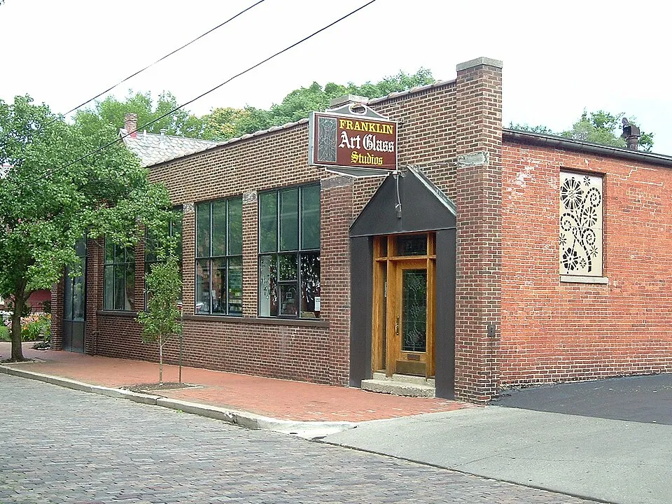 Exterior of Franklin Art Glass Studios, a single-story red brick commercial building on a brick-paved street corner in Columbus, Ohio. A hanging sign above the entrance reads "Franklin Art Glass Studios" in decorative lettering. The building features large industrial-style windows, a wooden front door with stained glass panels, a dark canopy over the entrance, and an ornate circular stained glass design embedded in the brick wall on the right side. Trees are visible in the background under an overcast sky.