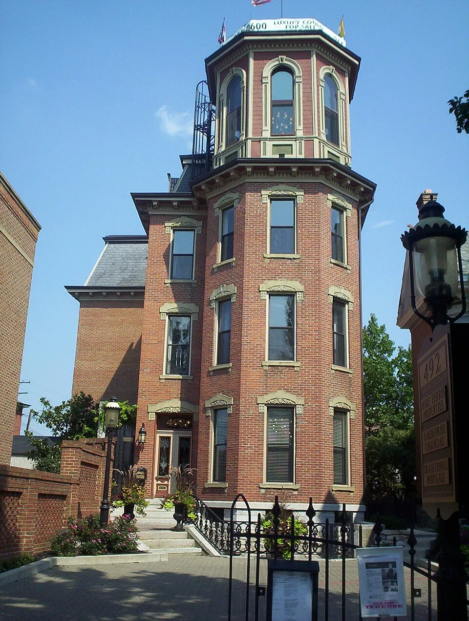 Exterior view of Schwartz Castle, a tall, narrow Victorian-era red brick building in German Village, Columbus, Ohio. The structure features an ornate octagonal cupola at the top with arched windows and small flags, decorative stone window surrounds, and a mansard-style roof on the adjoining wing. A black iron fence and gate line the front, with flower plantings along the entrance steps. A vintage-style street lamp is visible on the right side. A real estate sign near the top advertises luxury condos for sale.