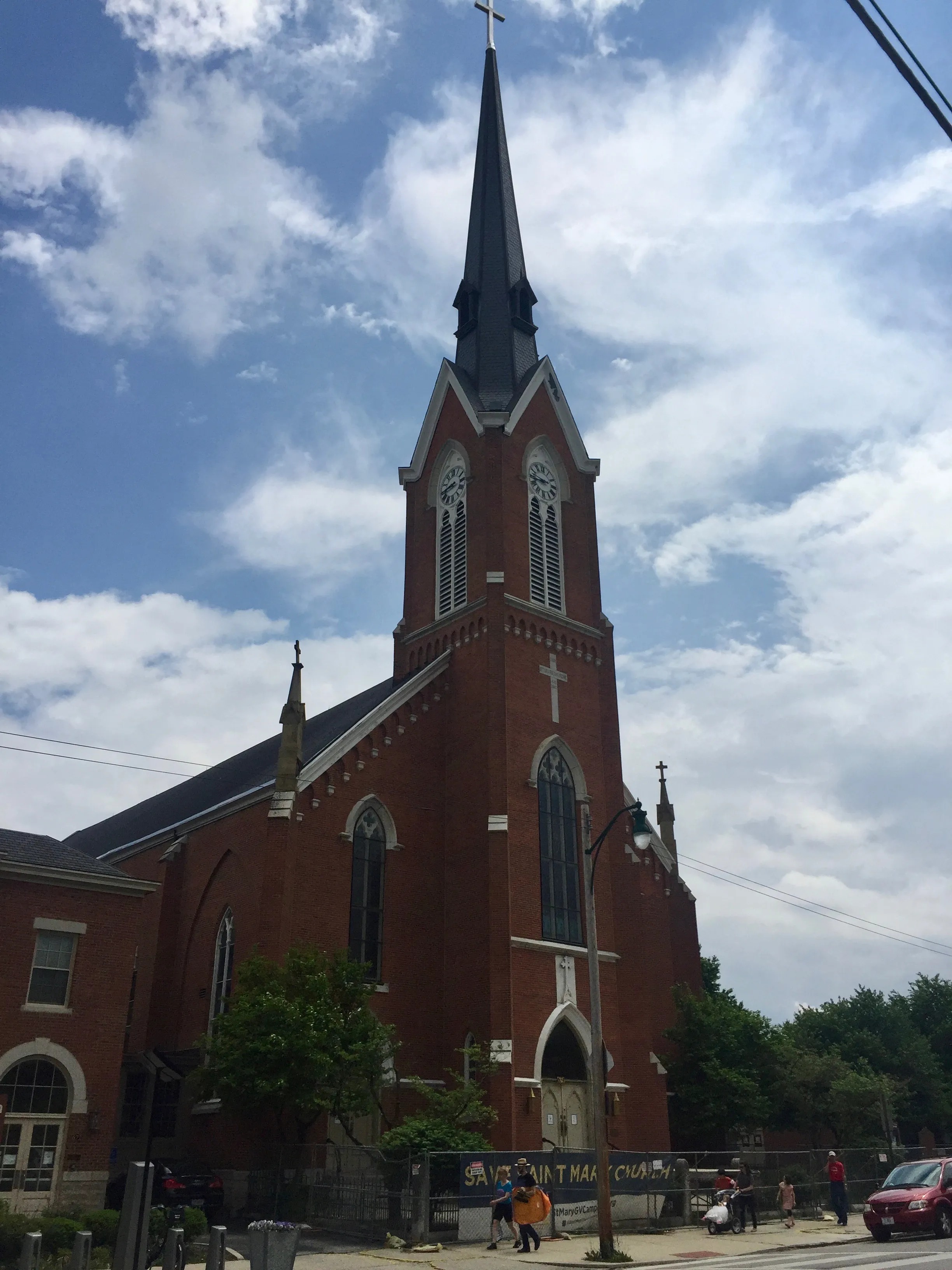 Exterior view of St. Mary Catholic Church in German Village, Columbus, Ohio. The red brick Gothic Revival-style church features a tall, dark steeple topped with a cross, a clock tower with two visible clock faces, pointed arch windows, and decorative white stone trim. A banner along the iron fence at street level reads "Save Saint Mary Church." Several people are visible on the sidewalk in front of the church. The photo is taken from street level looking upward against a partly cloudy blue sky.