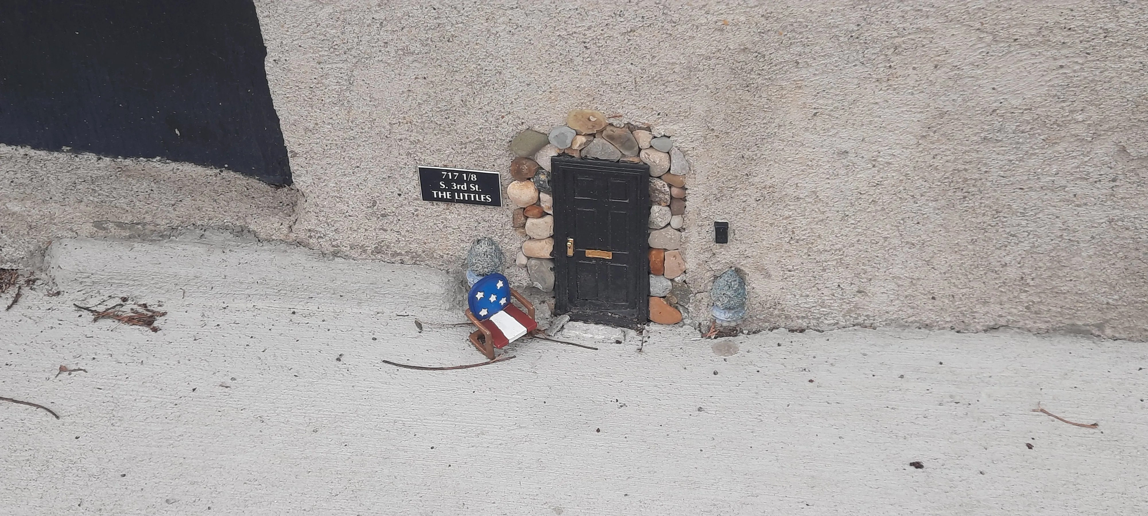 A tiny miniature black door built into the base of a textured concrete wall, framed by small river rocks. A small address plaque to the left reads "717 1/8 S. 3rd St. — THE LITTLES." In front of the door sits a miniature rocking chair painted in American flag colors (red, white, and blue with white stars), along with two small stone figurines flanking the entrance. The whimsical installation is set at ground level on a concrete sidewalk.