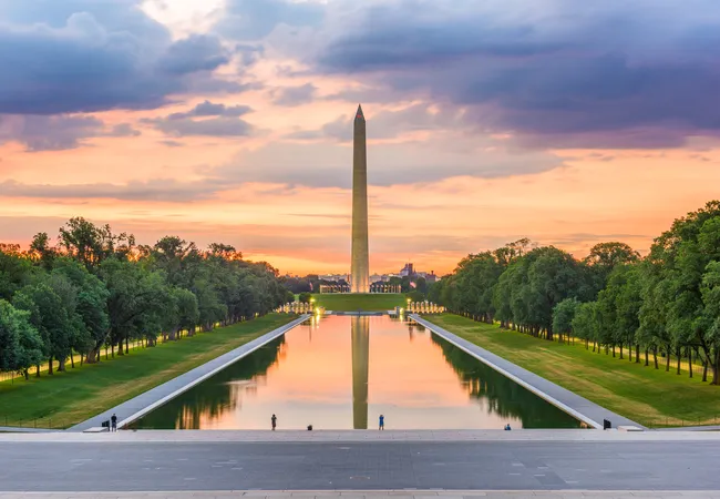 A symmetrical view from the steps of the Lincoln Memorial looking east along the National Mall in Washington, D.C., at sunset. The Lincoln Memorial Reflecting Pool stretches down the center, perfectly mirroring the Washington Monument and the vivid orange, pink, and purple sunset sky above. Rows of lush green trees line both sides of the mall, and the illuminated World War II Memorial is faintly visible midway. A handful of visitors stand along the pool's edge, dwarfed by the sweeping scale of the scene.