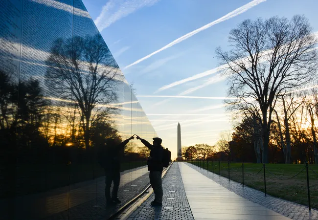 A silhouetted visitor stands with arms outstretched, touching the highly polished black granite surface of the Vietnam Veterans Memorial Wall in Washington, D.C., at sunset. The wall mirrors a vivid reflection of the golden sky, bare winter trees, and contrails streaking overhead. The Washington Monument is visible in the background between the trees, framed by the warm colors of dusk. The visitor's reflection appears in the wall alongside them, creating a powerful and intimate moment.