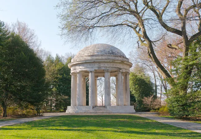 The District of Columbia World War I Memorial set within a park landscape in early spring in Washington, D.C. The circular neoclassical white marble domed rotunda stands on a raised stepped base surrounded by a lush green lawn. The open-air structure features tall fluted columns and a weathered domed roof, with the partial inscription "World War — A Memorial To" visible along the frieze. Mature trees — some bare, some evergreen — and trimmed shrubs frame the monument on both sides under a pale blue sky.