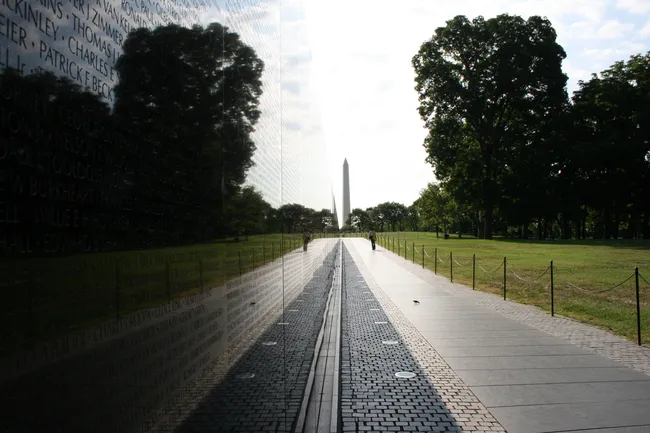 A perspective view along the Vietnam Veterans Memorial Wall on the National Mall in Washington, D.C. The highly polished black granite surface on the left reflects the surrounding trees, sky, and the distant Washington Monument, which is visible at the end of the path. Names of fallen service members are engraved across the wall in rows. A few visitors walk along the brick pathway beside the wall, bordered by a low chain fence on the right with lush green lawn and tall trees beyond. The image captures the wall's striking reflective quality and solemn scale.