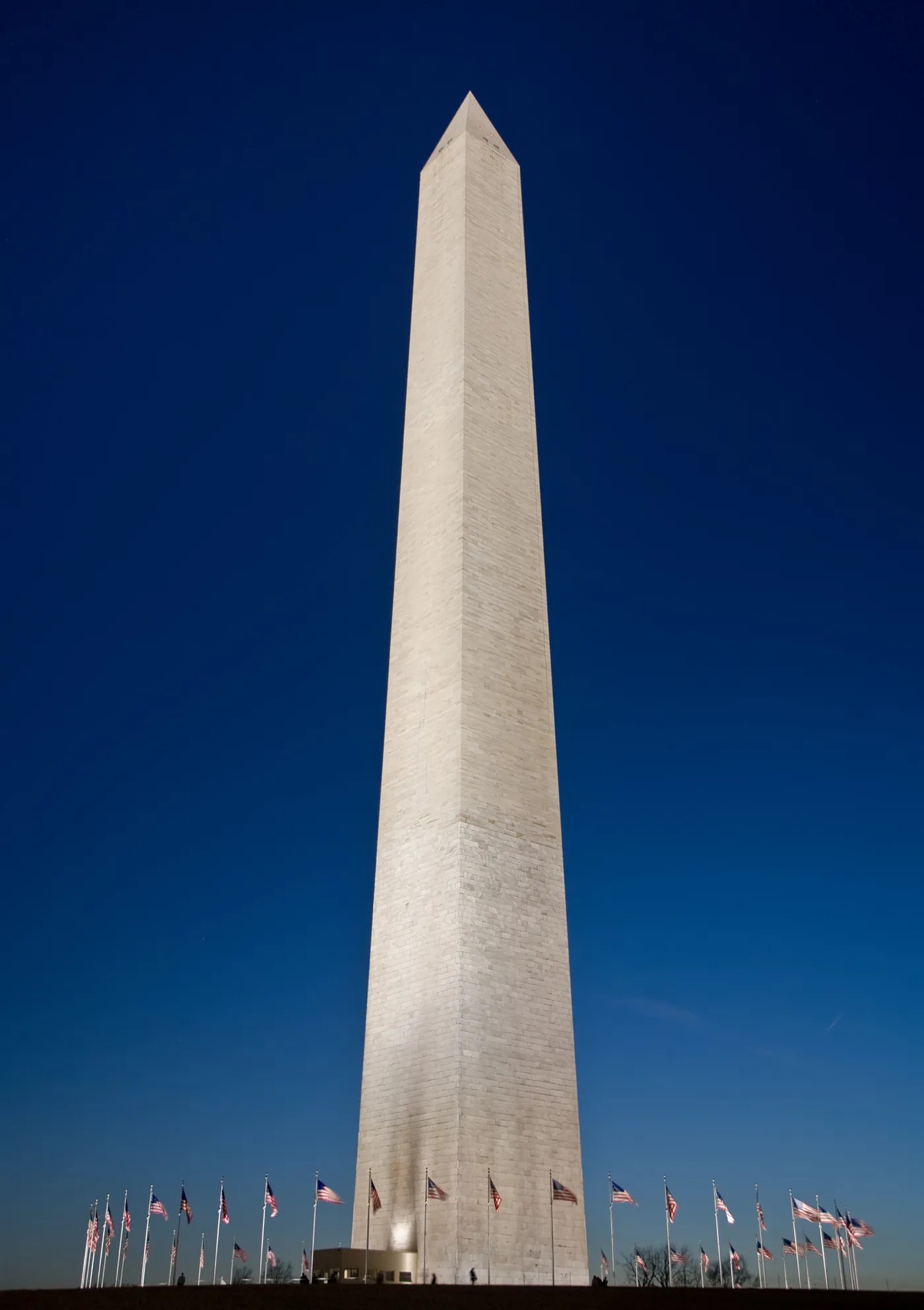 A dramatic low-angle view of the Washington Monument, a towering white marble obelisk, photographed at dusk against a deep, clear blue sky. The monument is illuminated by ground lighting, highlighting its pale stone surface. A ring of American flags on tall flagpoles encircles the base of the monument. Tiny silhouettes of visitors are faintly visible at ground level, emphasizing the monument's immense height. Washington, D.C.