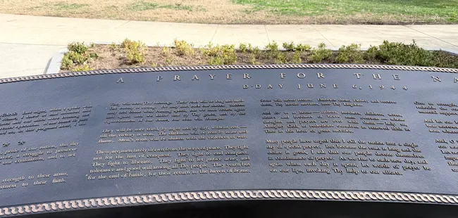 A curved dark bronze memorial plaque engraved with text titled "A Prayer for the N—" (partially cut off at the edge) and dated "D-Day June 6, 1944." The lengthy inscription contains the full text of President Franklin D. Roosevelt's prayer broadcast to the nation on the evening of the D-Day invasion. The plaque is set at an angle into a stone surface with a lawn and plantings visible in the background. This is part of a memorial in Washington, D.C.