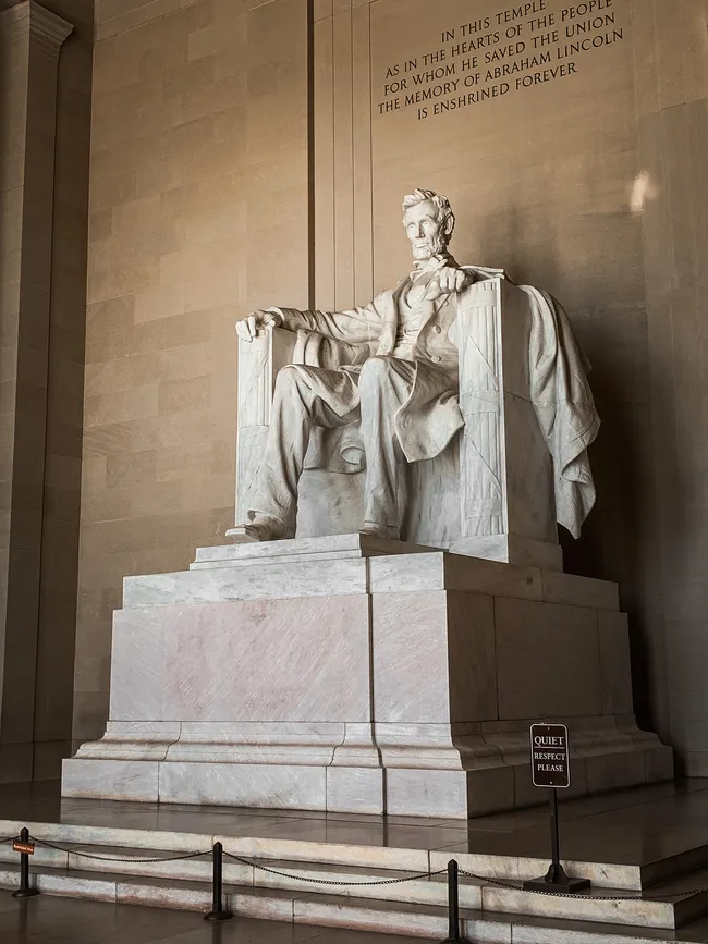 Interior view of the Lincoln Memorial in Washington, D.C., featuring the iconic seated marble statue of Abraham Lincoln sculpted by Daniel Chester French. Lincoln is depicted in a large throne-like chair, gazing solemnly forward with his hands resting on the armrests. The statue sits atop a multi-tiered marble pedestal. Inscribed on the wall above reads: "In this temple, as in the hearts of the people for whom he saved the Union, the memory of Abraham Lincoln is enshrined forever." A small "Quiet — Respect Please" sign and a rope barrier are visible at the base.