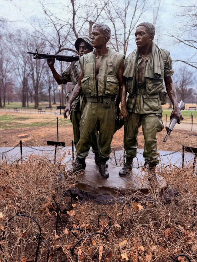 "The Three Soldiers," a bronze statue by Frederick Hart near the Vietnam Veterans Memorial on the National Mall in Washington, D.C. The sculpture depicts three young male soldiers in Vietnam-era combat gear — one holding an M16 rifle raised, another standing at center looking forward, and a third to the right holding a weapon at his side. The figures face toward the Memorial Wall. The statue is surrounded by a low chain barrier, dry winter leaves, and dormant brush, photographed on a gray, overcast day with bare trees in the background.