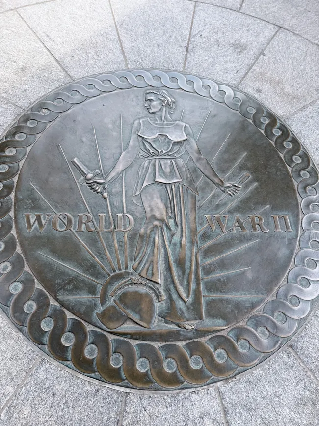 A circular bronze medallion set into the granite pavement at the National World War II Memorial in Washington, D.C. The relief depicts a classical female allegorical figure standing amid radiating sunbeams, holding a sword in one hand with a military helmet at her feet. The words "World War II" are inscribed on either side of the figure. The medallion is framed by a decorative rope-like border and surrounded by gray granite paving stones.