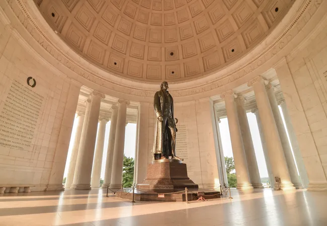 Interior view of the Thomas Jefferson Memorial in Washington, D.C., looking up at the elegant coffered dome ceiling. The tall bronze statue of Thomas Jefferson stands on a red granite pedestal at the center of the circular chamber, surrounded by tall Ionic columns that open to views of trees and sky beyond. Inscribed text from Jefferson's writings is visible on the curved interior walls to the left. Warm, diffused light fills the space, reflecting off the polished marble floor.