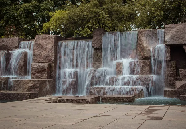 A long-exposure photograph of the multi-tiered waterfall feature at the Franklin Delano Roosevelt Memorial in Washington, D.C. Water cascades smoothly down several levels of large, rough-cut red granite blocks, creating a silky, misty effect as it collects in a shallow reflecting pool at the base. A broad granite plaza stretches across the foreground, and dense green trees rise above the granite walls in the background under an overcast sky.