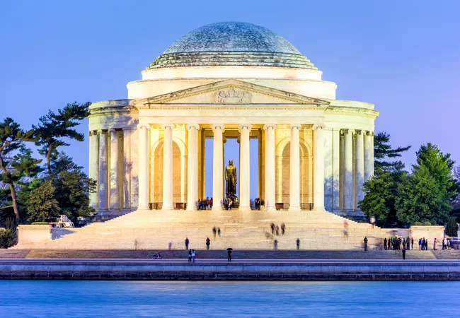 The Thomas Jefferson Memorial illuminated at dusk, viewed from across the Tidal Basin in Washington, D.C. The neoclassical circular marble structure glows with warm golden light, highlighting its tall Ionic columns, triangular pediment, and large patinated dome against a deep blue twilight sky. The bronze statue of Thomas Jefferson is visible standing within the open rotunda. Visitors are scattered on the broad steps and along the water's edge, with long-exposure motion blur suggesting a lively evening scene. Pine trees frame both sides of the memorial.
