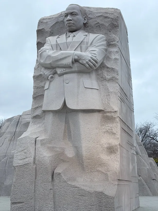 A close-up view of the Martin Luther King Jr. Memorial in Washington, D.C. The massive light gray granite sculpture depicts Dr. King emerging from a large rough-hewn stone block, arms folded across his chest, wearing a suit and tie, with a solemn and resolute expression. The figure appears to rise out of the "Mountain of Despair," referencing his iconic speech. Bare trees and an overcast gray sky are visible in the background.