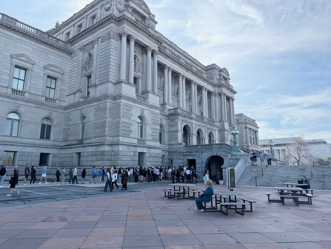 The exterior of the Library of Congress Thomas Jefferson Building in Washington, D.C., photographed from the plaza level. The grand Beaux-Arts granite facade features tall Corinthian columns, ornate sculptural reliefs, and arched entryways. A long queue of visitors stretches along the base of the building waiting to enter. Several people sit at picnic tables in the foreground plaza, and additional visitors are visible on the steps to the right. The scene is set under a partly cloudy sky in the early evening.