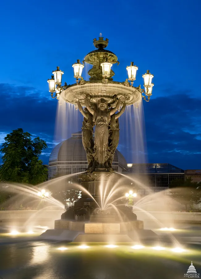 The Bartholdi Fountain photographed at dusk in Bartholdi Park near the U.S. Botanic Garden in Washington, D.C. The ornate cast-iron fountain, designed by Frédéric Auguste Bartholdi, features three large classical female figures holding aloft a wide basin from which water cascades down. Multiple illuminated gas-style lanterns crown the upper tier, glowing warmly against a deep blue twilight sky. Water jets spray outward at the base, lit from below by underwater lights. The dome of the U.S. Botanic Garden Conservatory is visible in the background.