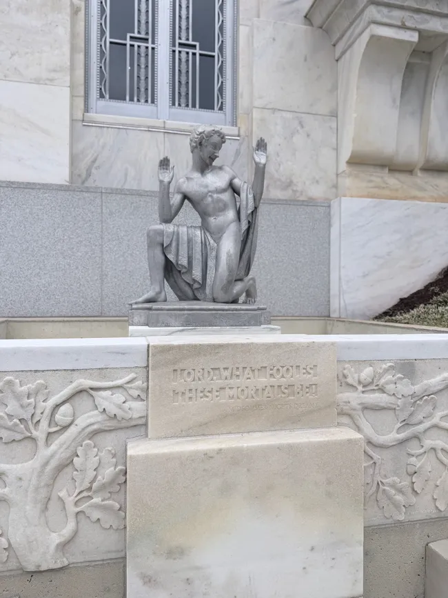 A stone sculpture of a kneeling male figure with both arms raised, draped in cloth, set on a pedestal against the white marble exterior of a grand building. Below the figure, a stone panel is carved with oak leaf and acorn relief decorations and the inscription: "Lord, what fools these mortals be!" — a quote from Shakespeare's A Midsummer Night's Dream. The setting appears to be the exterior of the Folger Shakespeare Library in Washington, D.C.