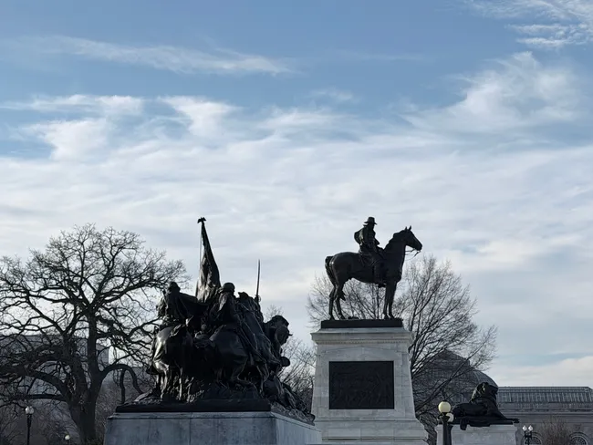 A low-angle silhouette view of the Ulysses S. Grant Memorial at the base of Capitol Hill in Washington, D.C. On the right, a bronze equestrian statue of General Grant sits atop a tall rectangular stone pedestal. To the left, a dramatic group sculpture depicts Civil War cavalry soldiers in chaotic motion, with a flag bearer and soldiers on horseback. Bare winter trees frame the scene against a pale blue sky streaked with wispy clouds. The glass dome of the U.S. Botanic Garden Conservatory is faintly visible in the background.