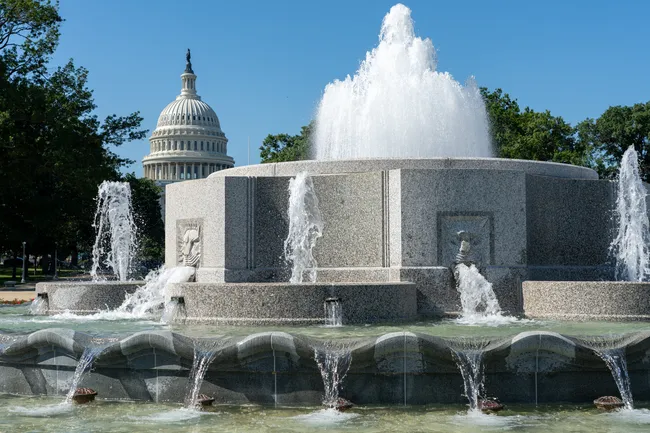 The Senate Fountain on the grounds of the U.S. Capitol in Washington, D.C., photographed on a sunny summer day. A powerful central water jet shoots high into the air from a large circular granite fountain structure, with multiple smaller jets cascading outward from the sides. Stone relief carvings of faces are visible on the fountain's exterior walls. The iconic white dome of the U.S. Capitol building rises prominently in the background, framed by lush green trees under a clear blue sky.