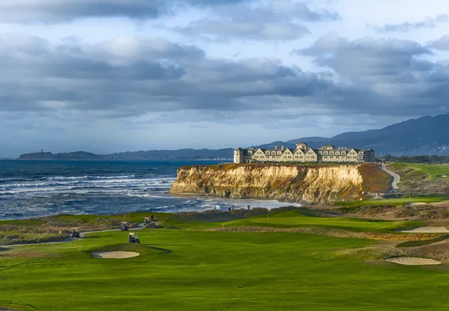 A coastal golf course stretches along the shoreline, with green fairways overlooking ocean waves and a large cliffside building perched above rugged bluffs under a cloudy sky.