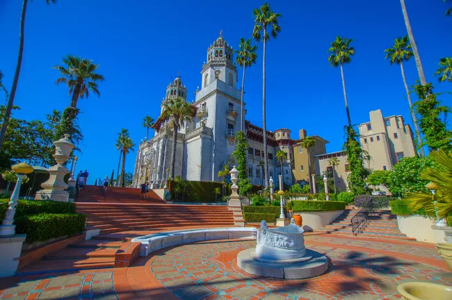 Hearst Castle rises above a grand terraced courtyard lined with palm trees, featuring ornate twin towers, sweeping staircases, and Mediterranean-style architecture beneath a clear blue sky.