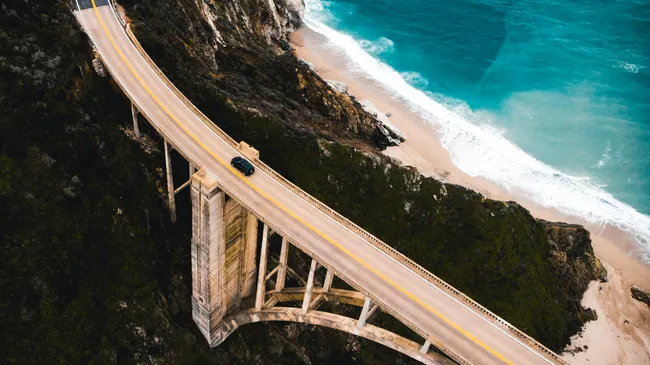 An aerial view of Bixby Creek Bridge carrying a single vehicle across a dramatic coastal canyon, with turquoise waves rolling onto a sandy beach below and steep cliffs lining the Big Sur shoreline.